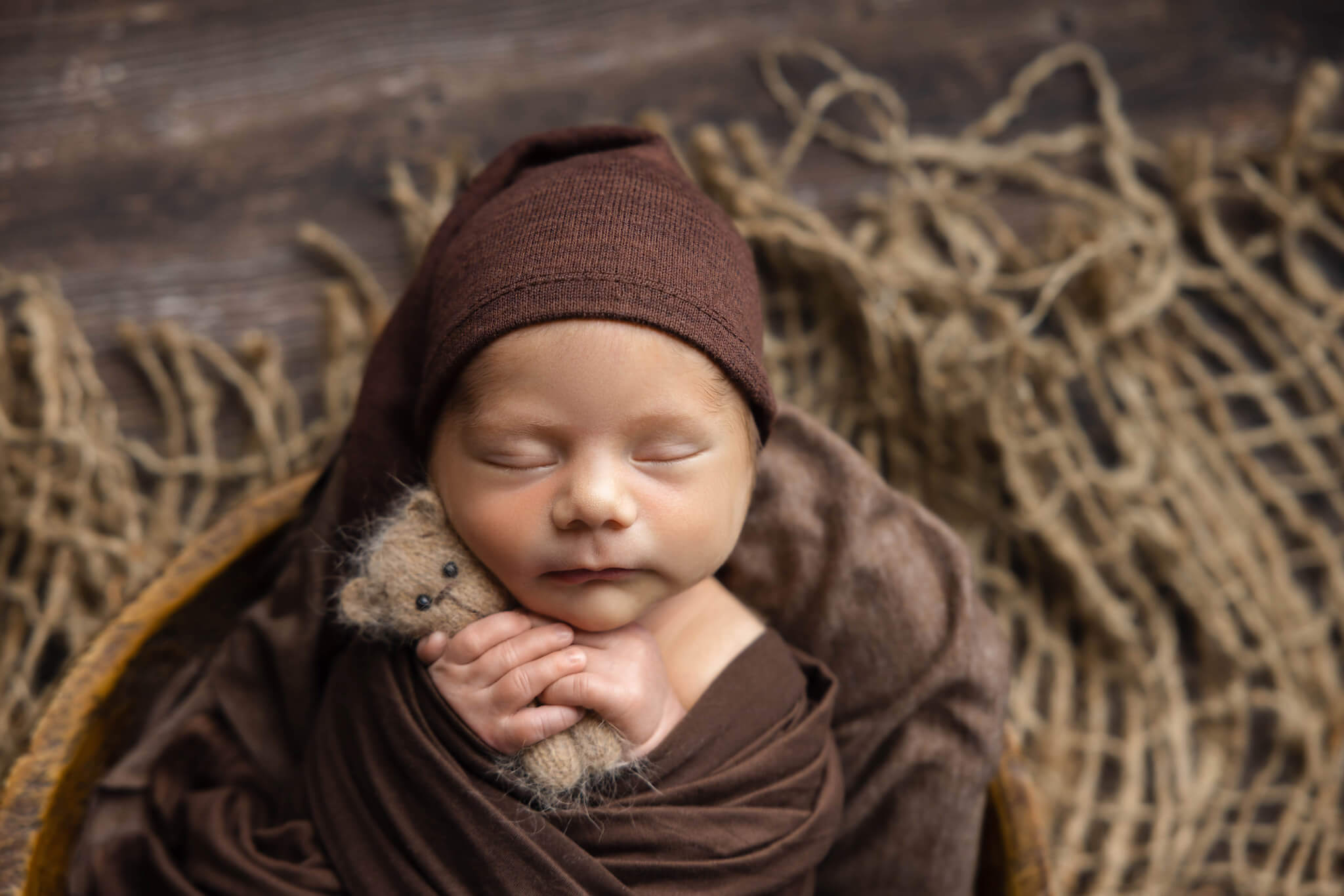 close up of a sleeping newborn baby in a brown wrap with a brown sleepy cap holding a tiny teddy bear