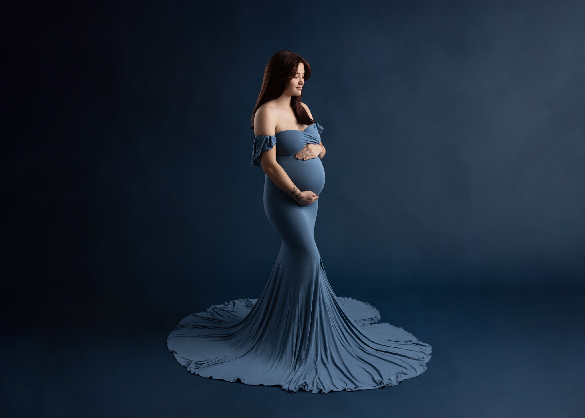 pregnant woman in an off the shoulder blue fitted dress standing holding her bump looking at the ground