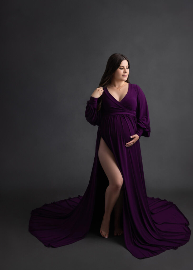 pregnant woman in a vibrant purple flowy dress standing in front of a dark grey backdrop holding her belly with one hand and twirling her hair with the other