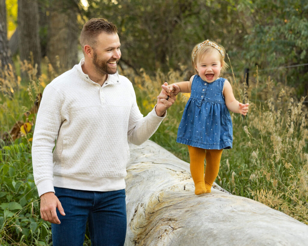 young dad in a sweater and jeans holding his young daughter's hand as she walks along a long smiling in City Park Pueblo, CO