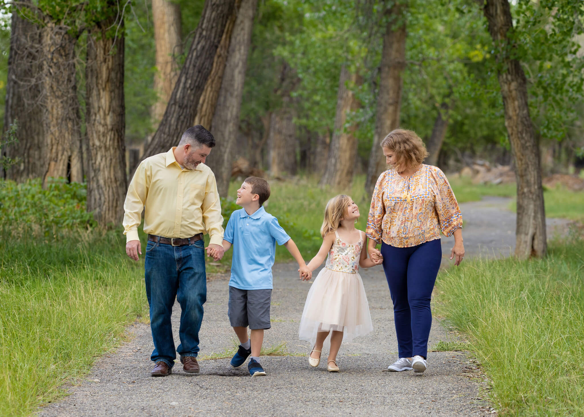 a family of 4 holding hands walking along a paved path in a forested area showing a thing to do in Pueblo with kids