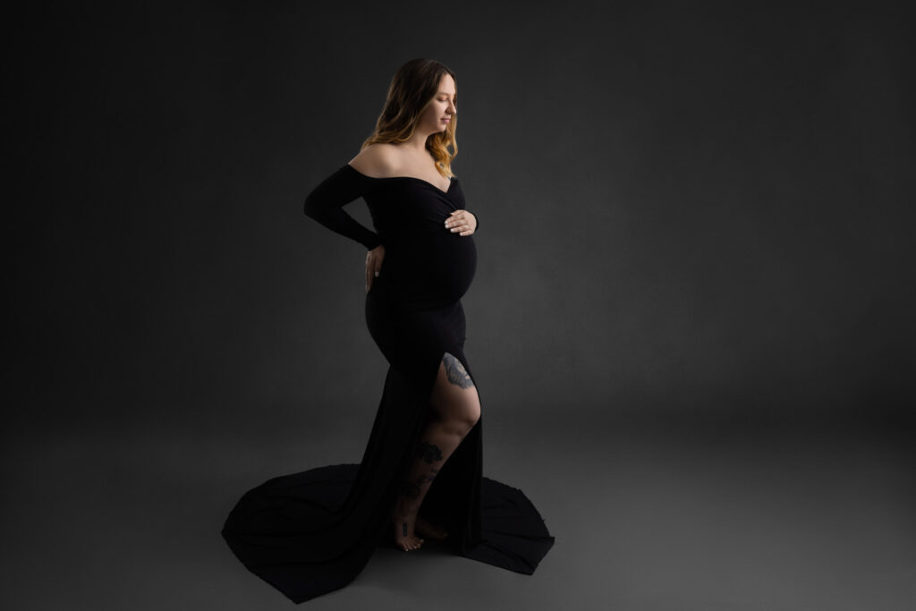 young pregnant mom wearing an off the shoulder black dress with one hand on her back and the other on the top of her belly standing on a grey background