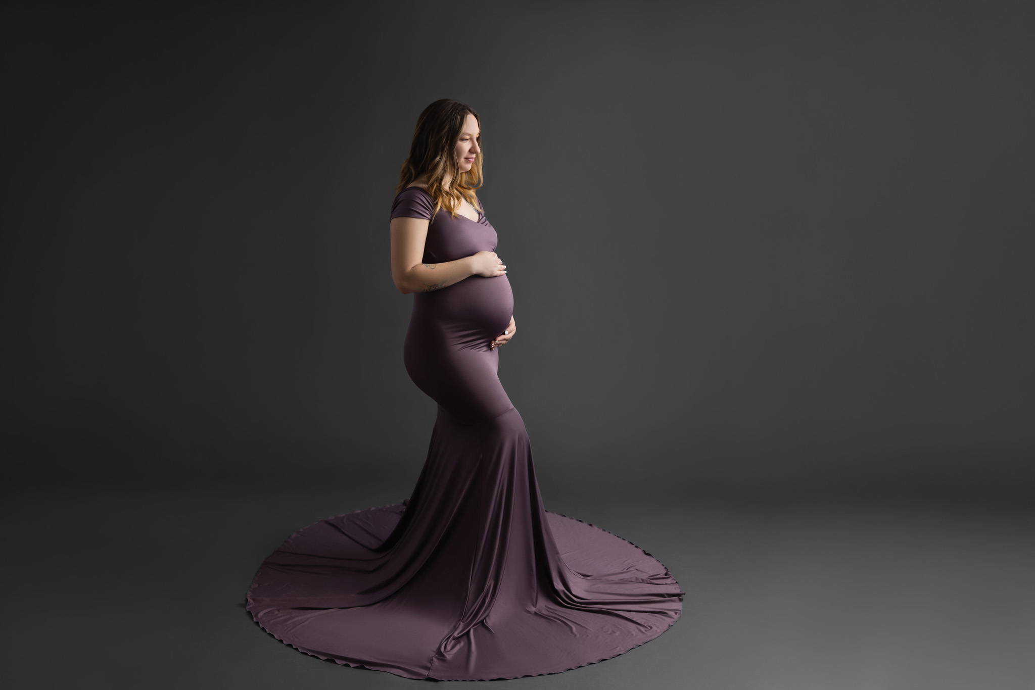 young pregnant mom wearing a purple dress with a long skirt with one hand on her back and the other on the top of her belly standing on a grey background