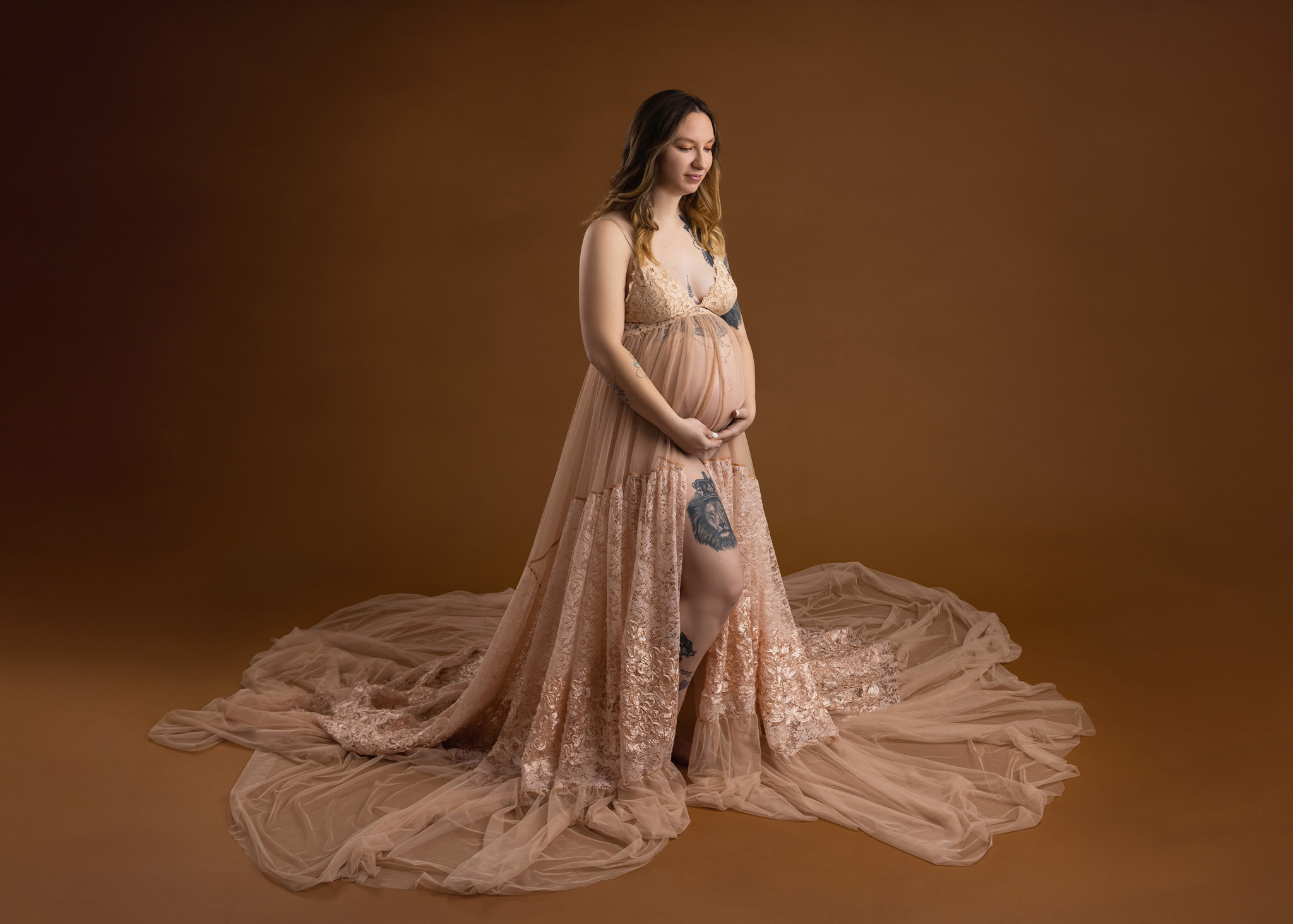 pregnant mom holding her belly with her gace downward wearing a lacy flowy tan dress standing on a brown backdrop