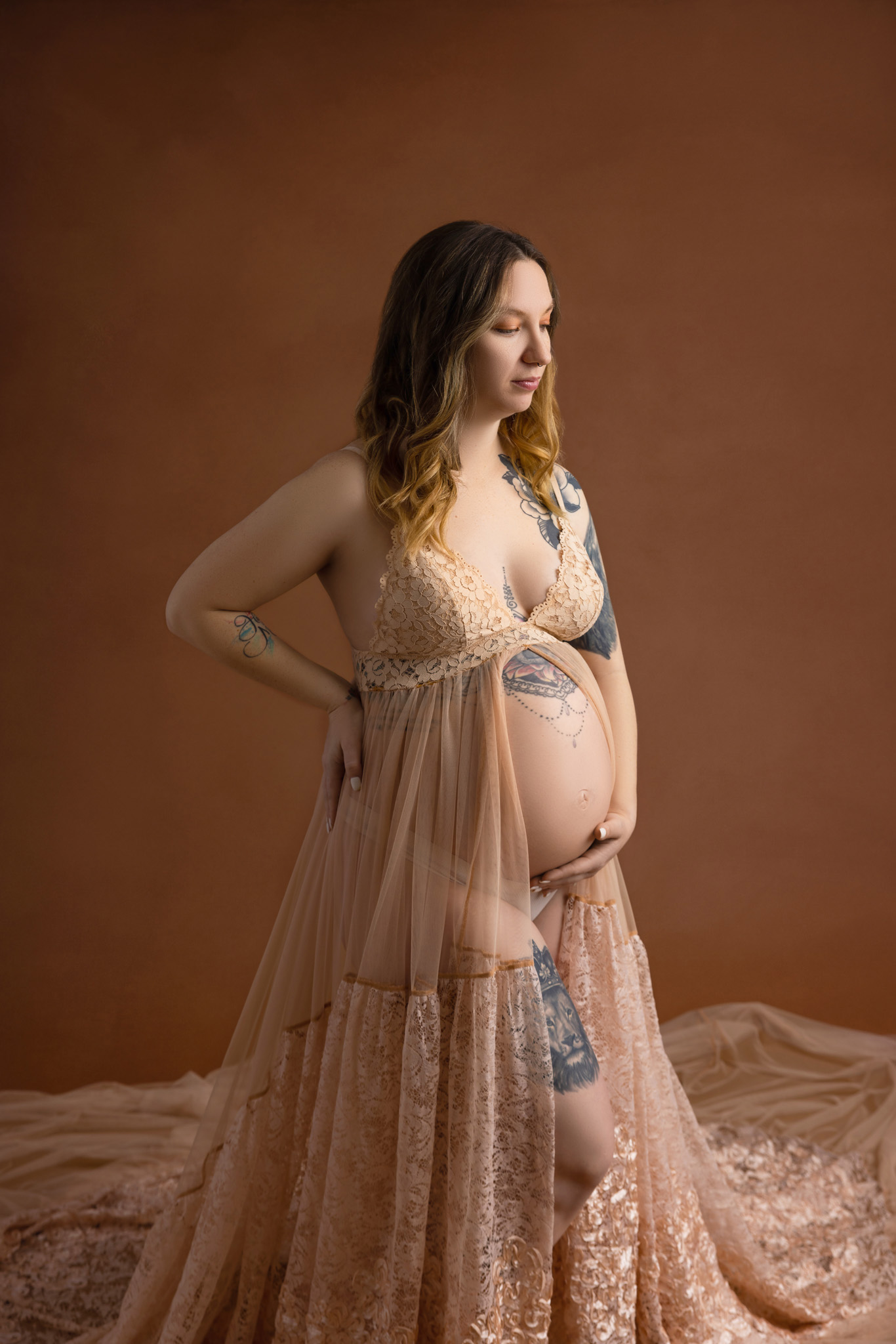 pregnant mom with one hand on her back and the other on her belly with her gace downward wearing a lacy flowy tan dress standing on a brown backdrop