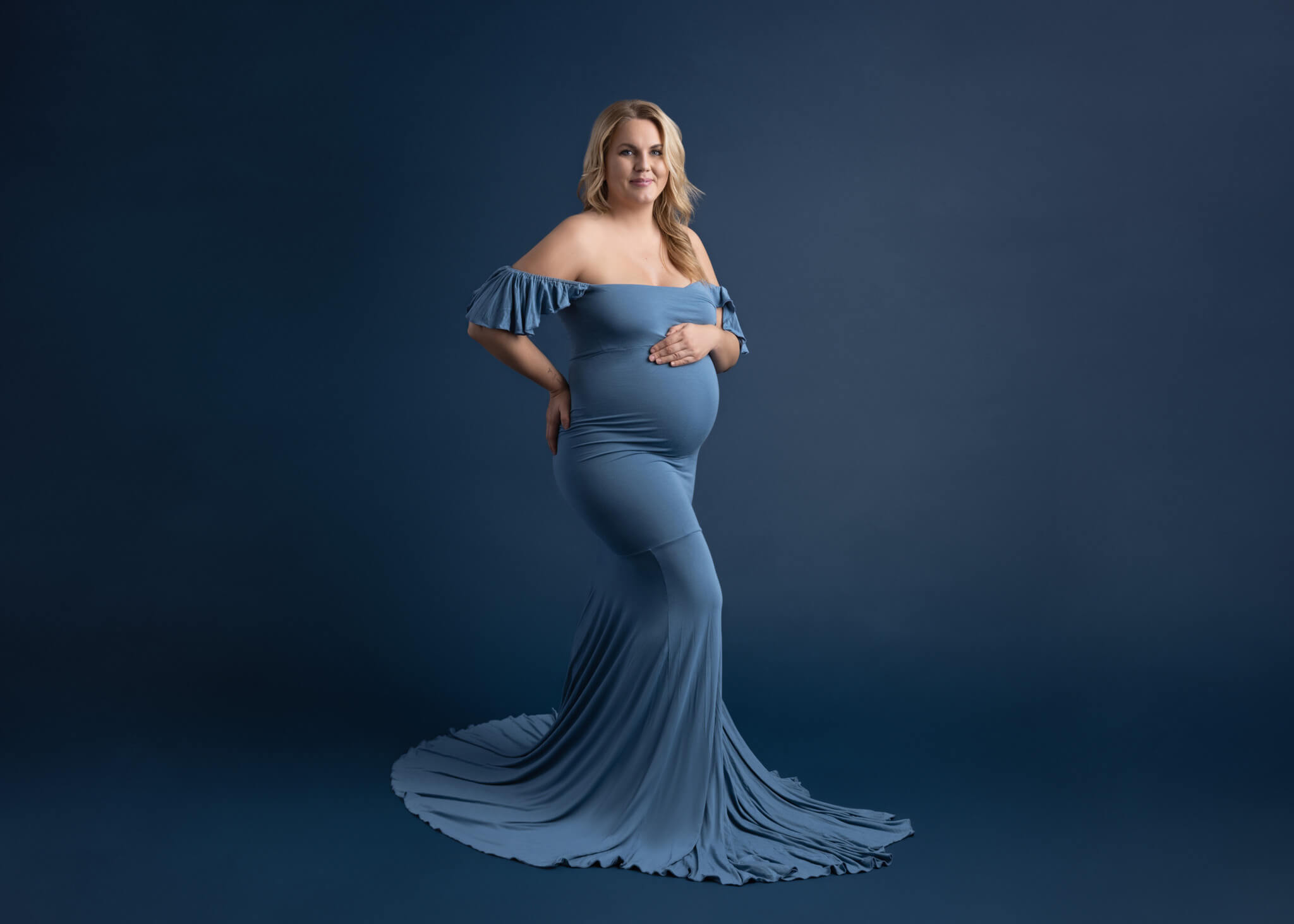 a pregnant mom wearing an off the shoulder blue fited dress standing holding her belly in front of a blue backdrop