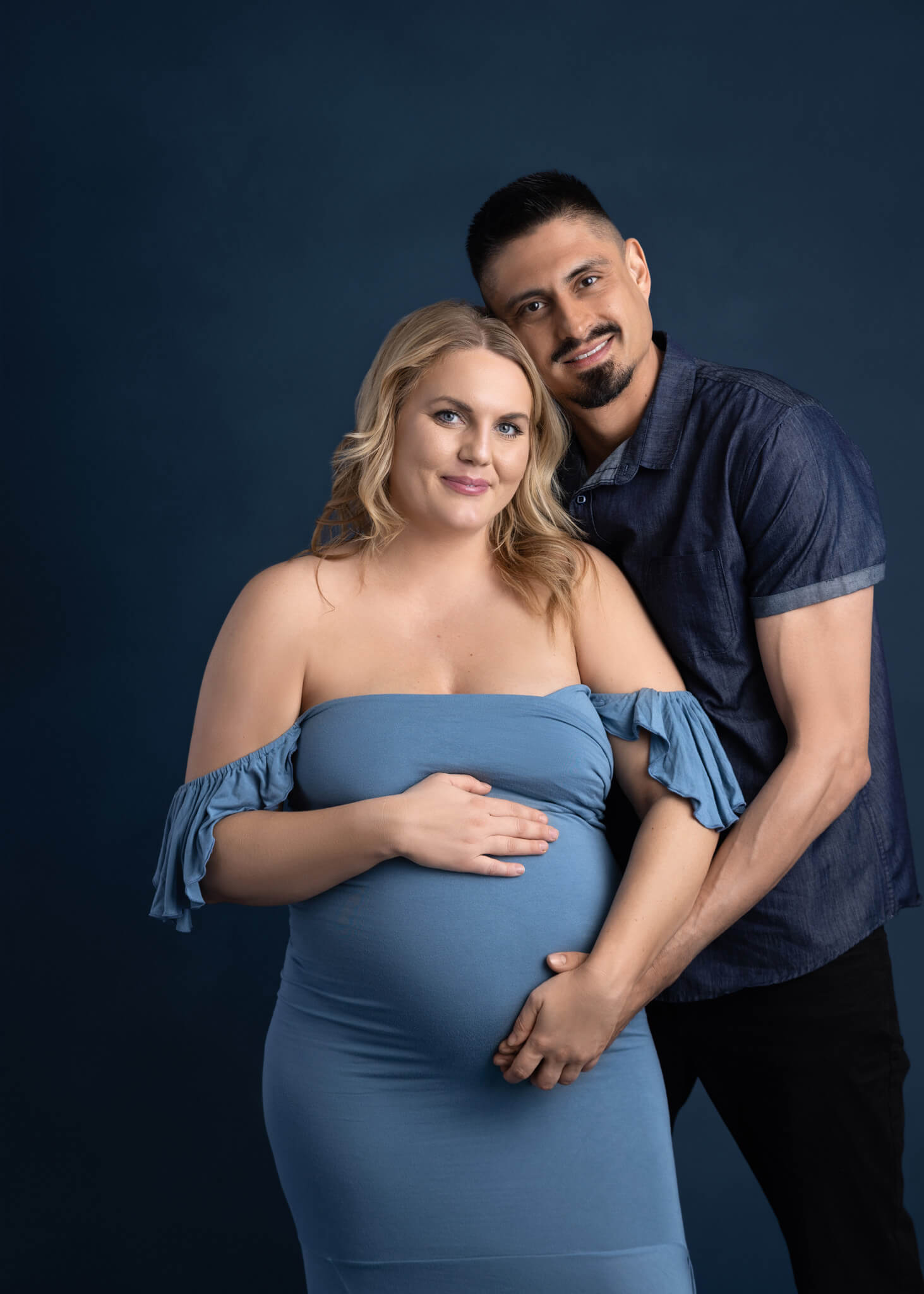 a pregnant mom in a fitted blue off the shoulder dress and her husband hugging close while both have a hand on her belly