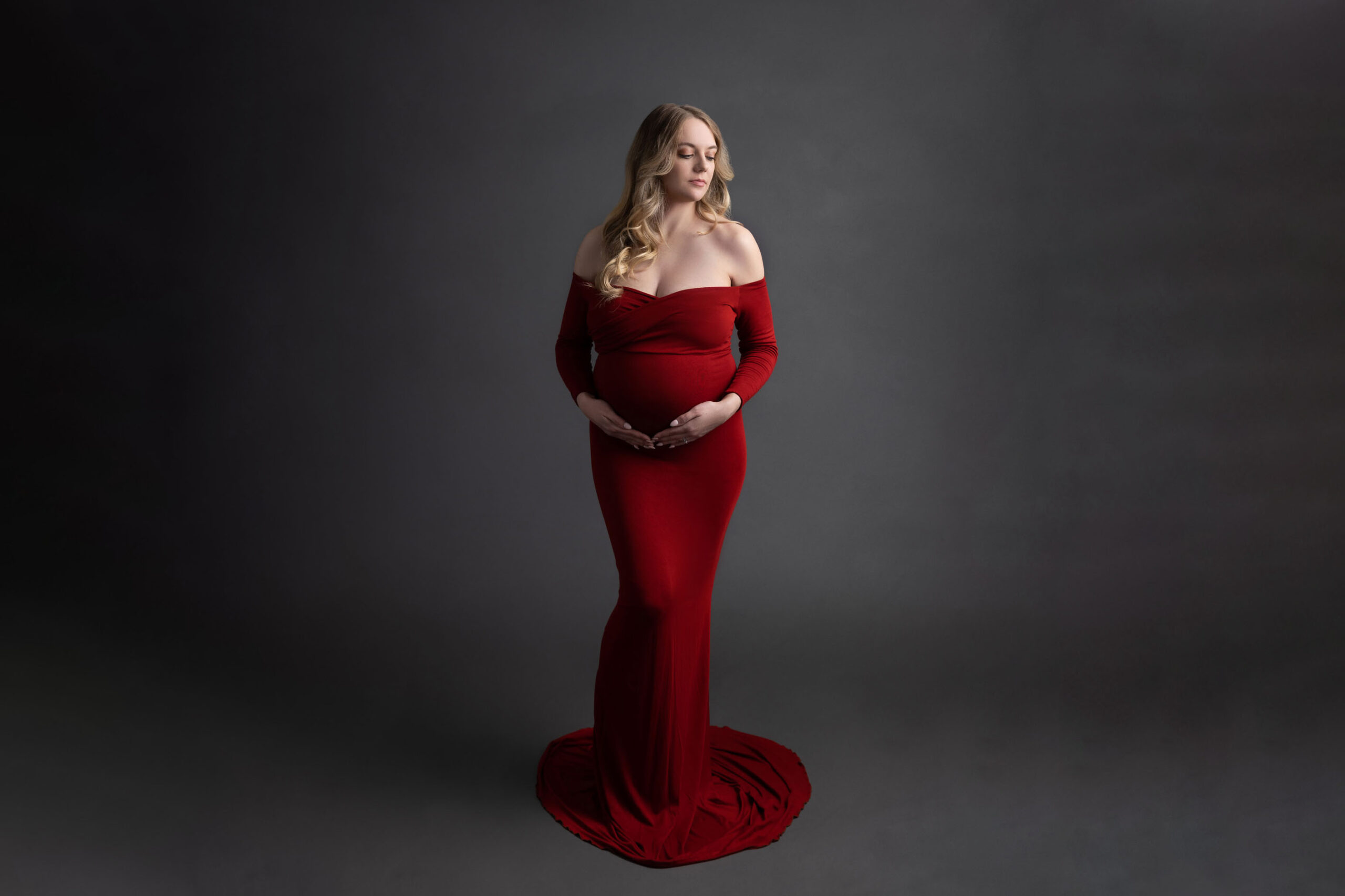pregnant mom wiht blond hair in an off the shoulder red dress fitted dress holding her belly with both hands underneath staring off to the right standing in front a grey backdrop