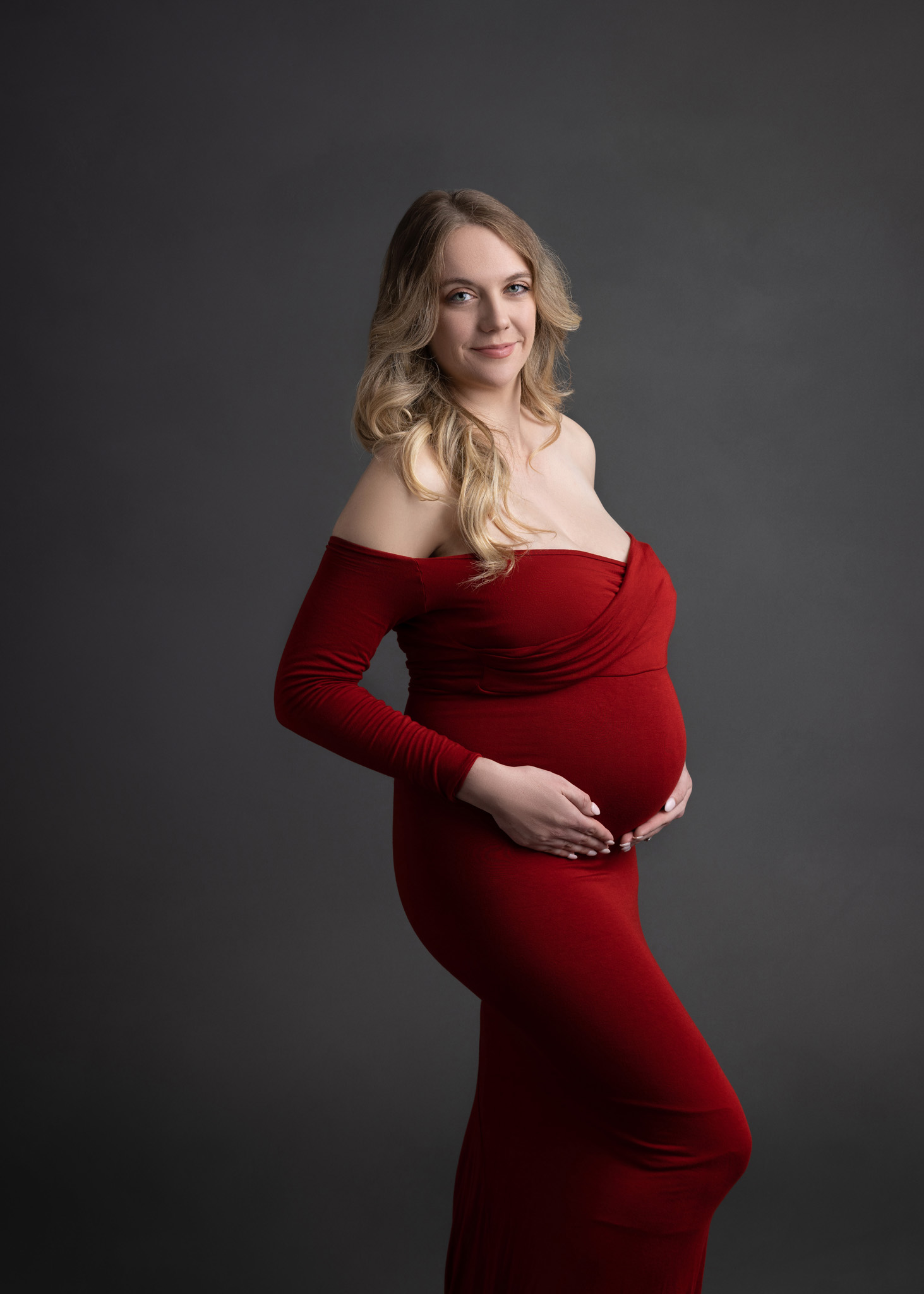 side profile of a pregnant mom in a fitted off the shoulder red dress cradling her belly with both hands standing in front of a grey backdrop