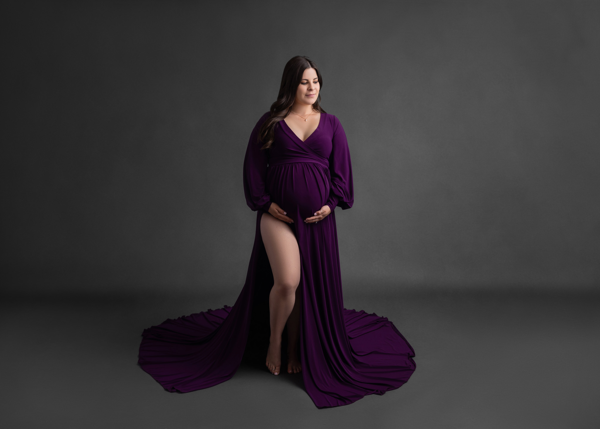 pregnant woman in a vibrant purple flowy dress standing in front of a dark grey backdrop holding her belly with both hands looking off to the distance