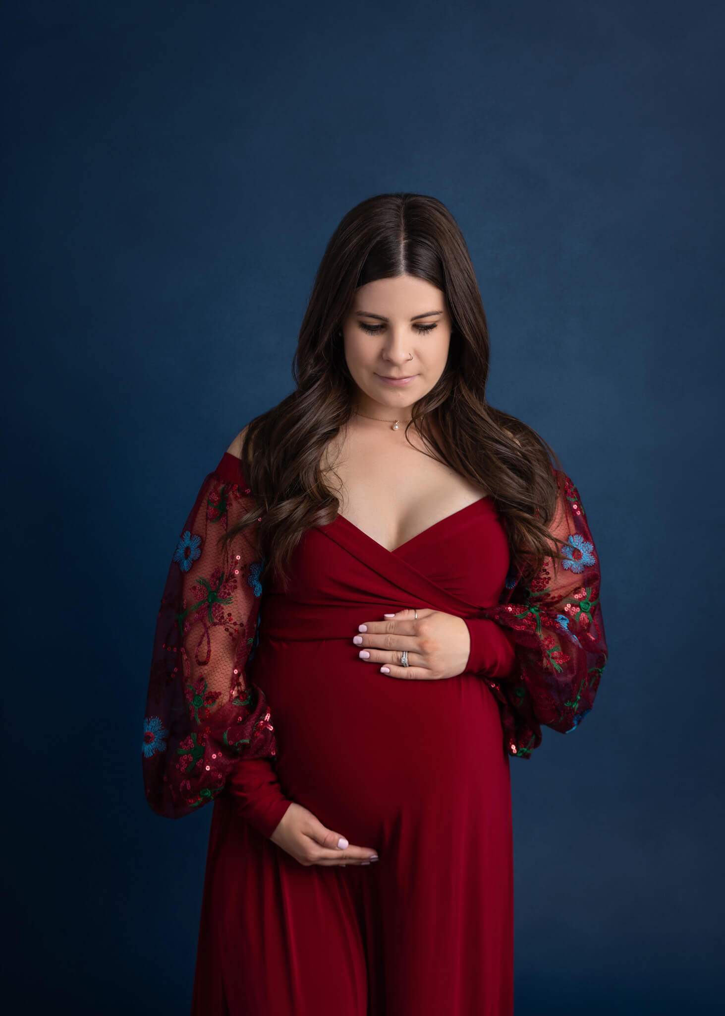 pregnant mom with long brown hair wearing a red dress looking down and holding her pregnant belly
