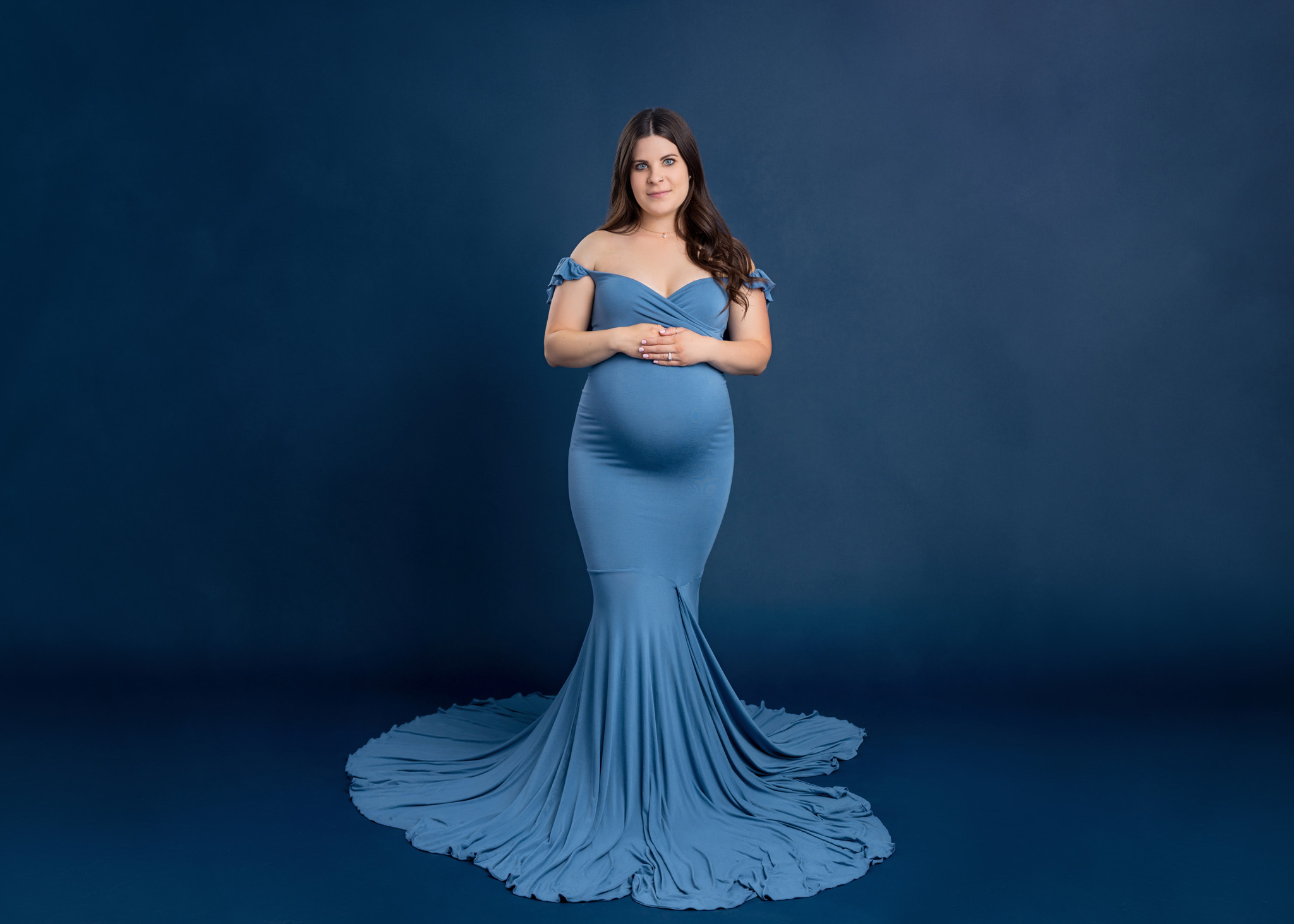 pregnant women in light blue off the shoulder dress standing in front of a cblue backdrop holding her pregnant belly gently gazing at the camera