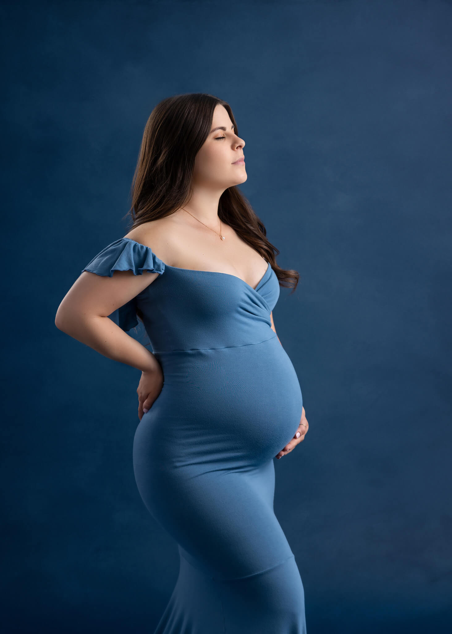 side profile of a pregnant woman in fitted light blue off the shoulder dress standing in front of a blue backdrop holding her pregnant belly 