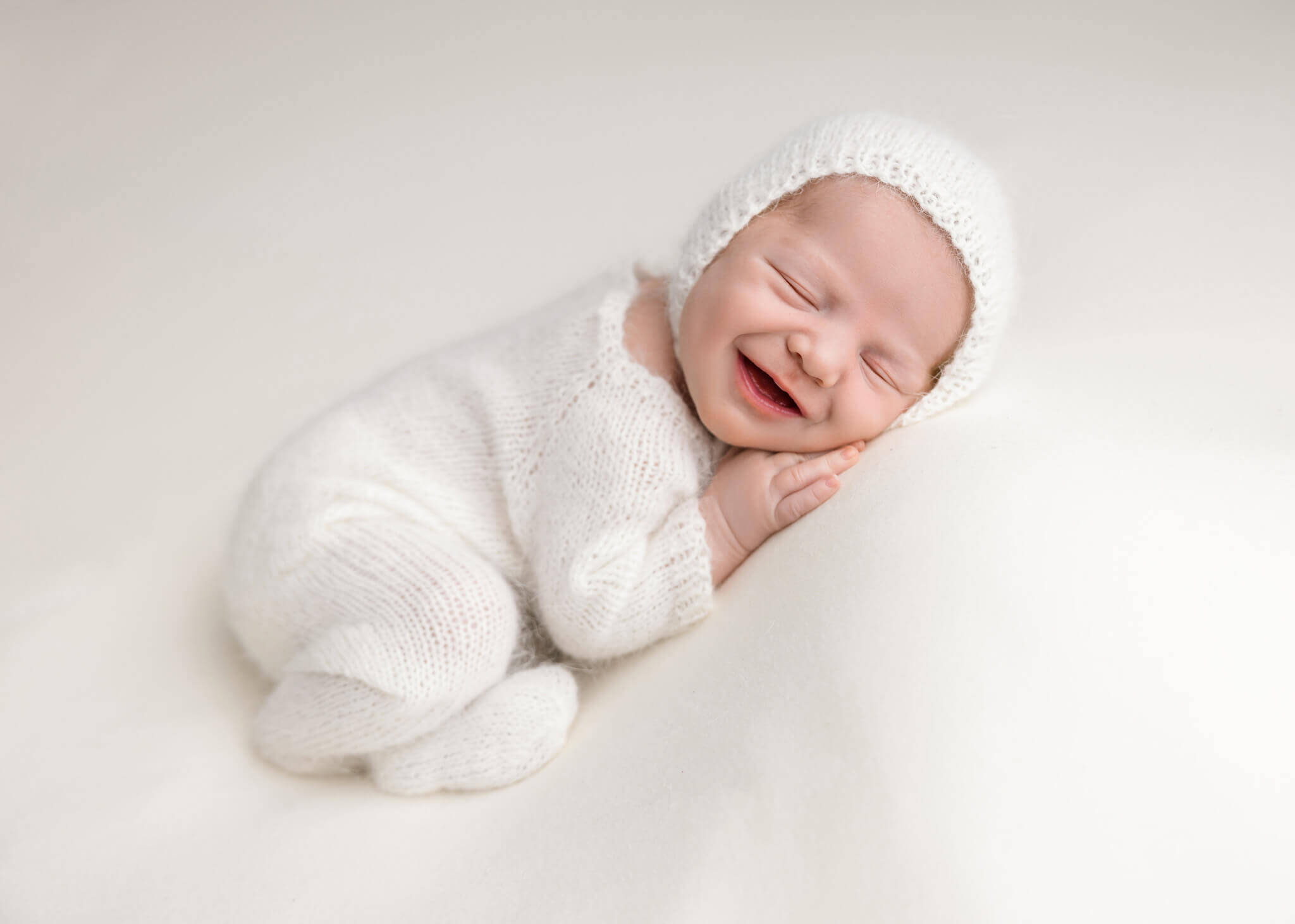 newborn baby in white romper with a white bonnet sleeping on a her side on a white blanket