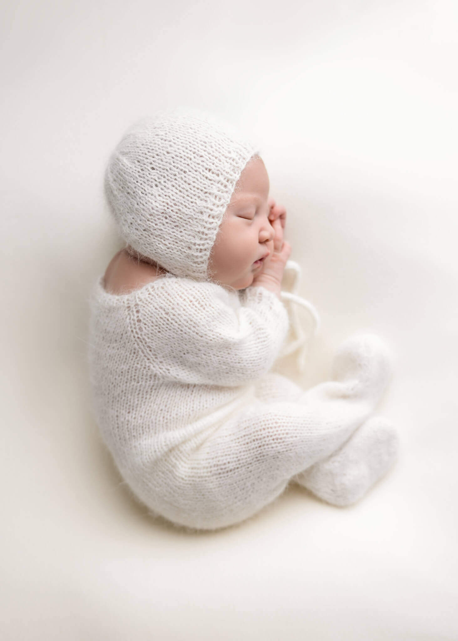 newborn baby in white romper with a white bonnet sleeping on a her side on a white blanket