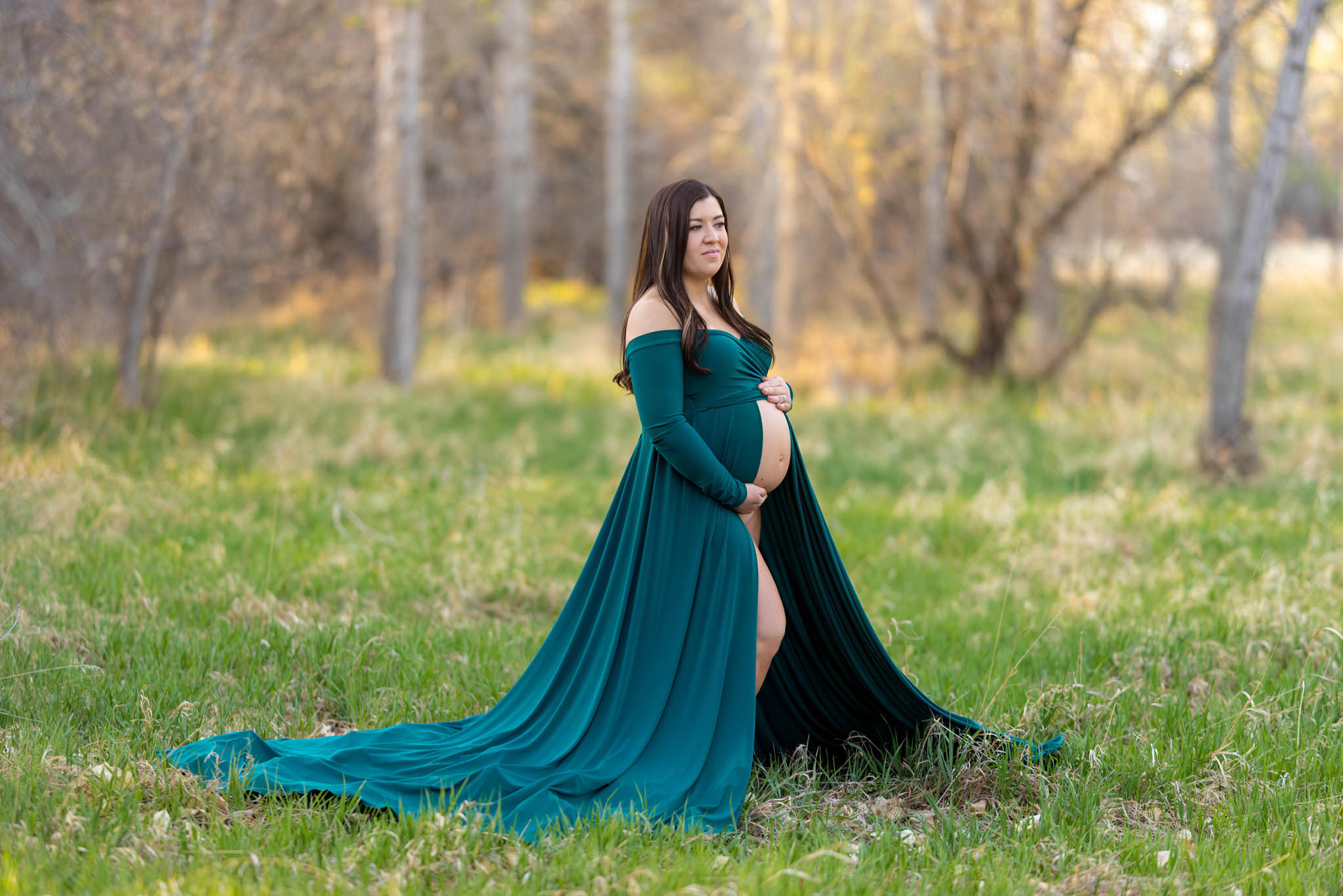 a pregnant mom in an off the shoulder green flowy dress holding her belly standing in a field in City Park in Pueblo CO