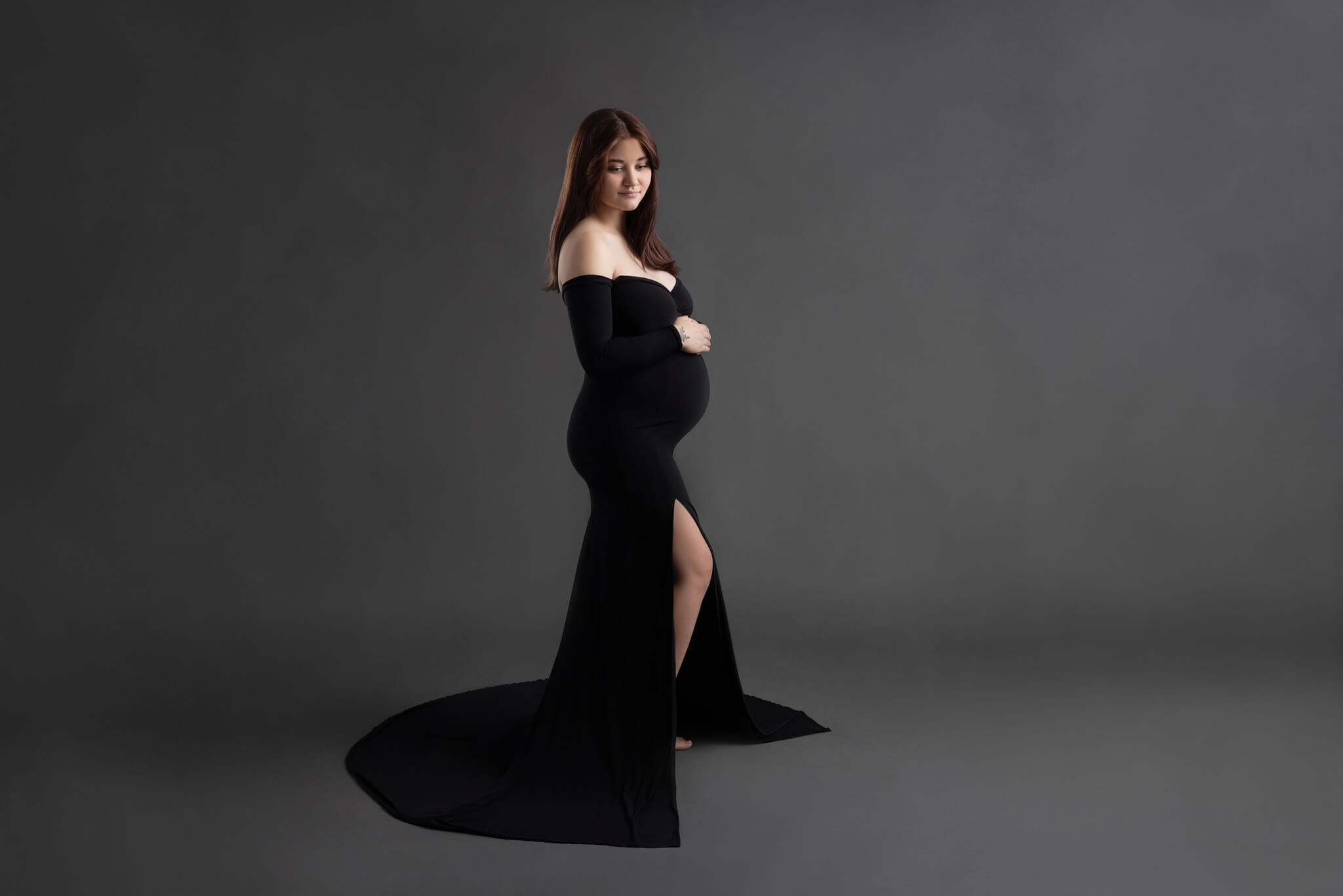 a side profile of a pregnant mom in an off the shoulder fitted dress standing against a grey backdrop with her hands resting on her belly