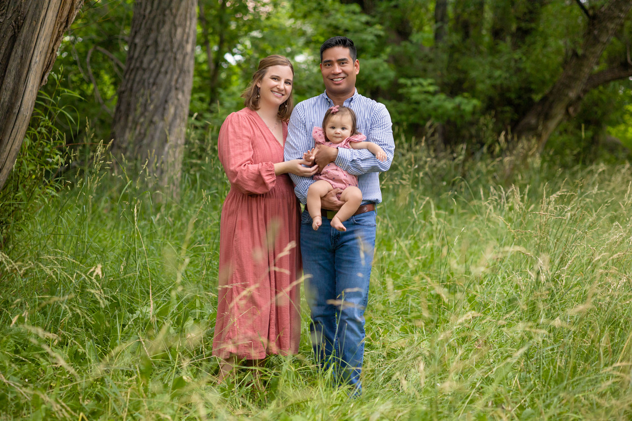 mom and dad holding young baby daughter standing together in a grassy field under trees in Pueblo CO