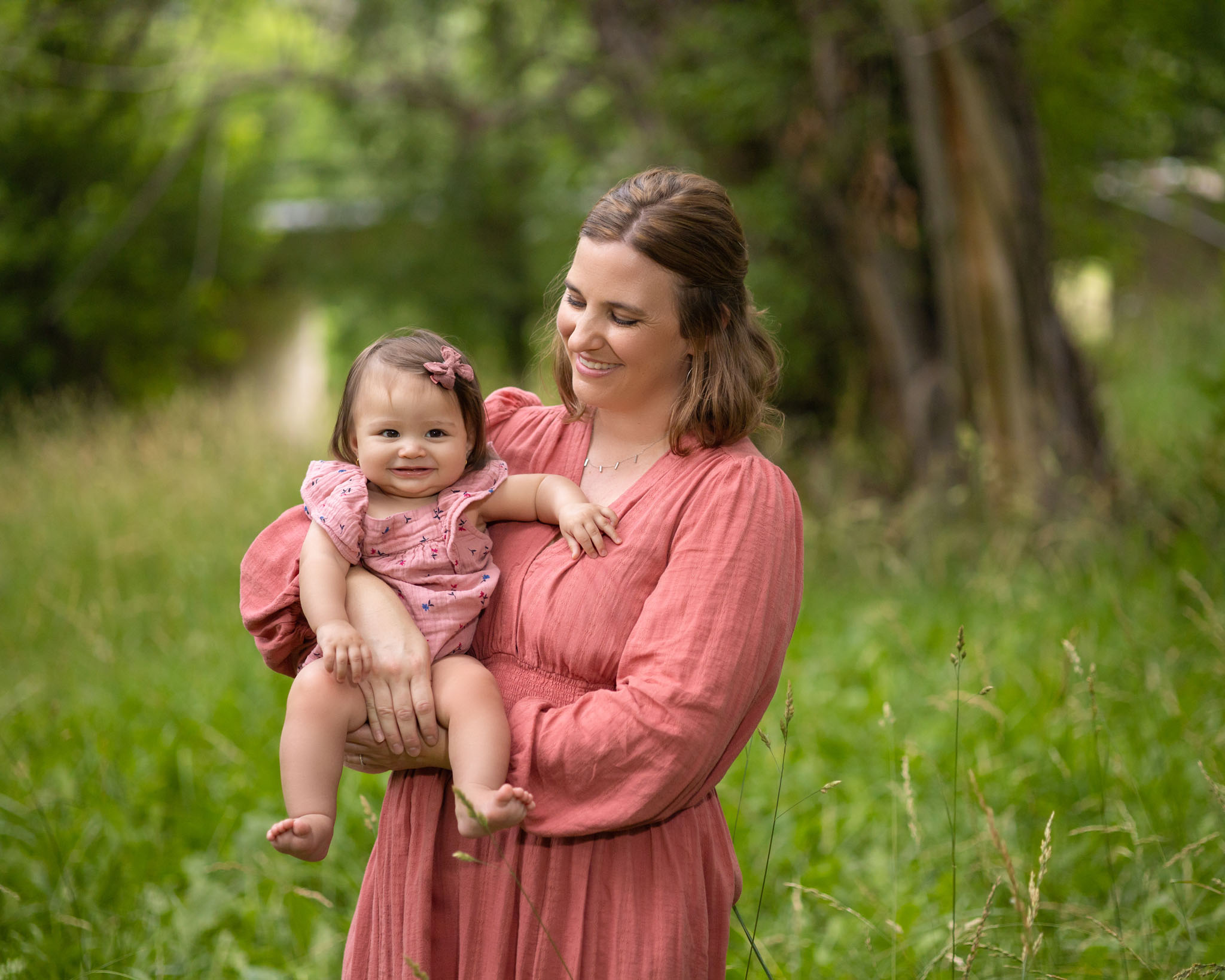 mom and baby daughter in pink outfits standing in a grassy field under trees in Pueblo CO