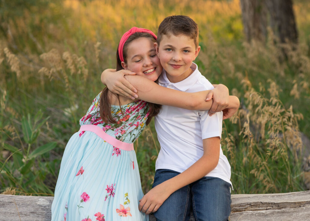 elementary aged sister hugging her brother with a big smile on her face sitting on a log in City Park Pueblo CO with golden tall grass around them