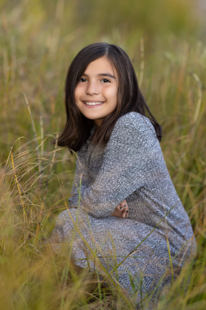 young brunette girl crouching in tall brown grasses at City Park in Pueblo