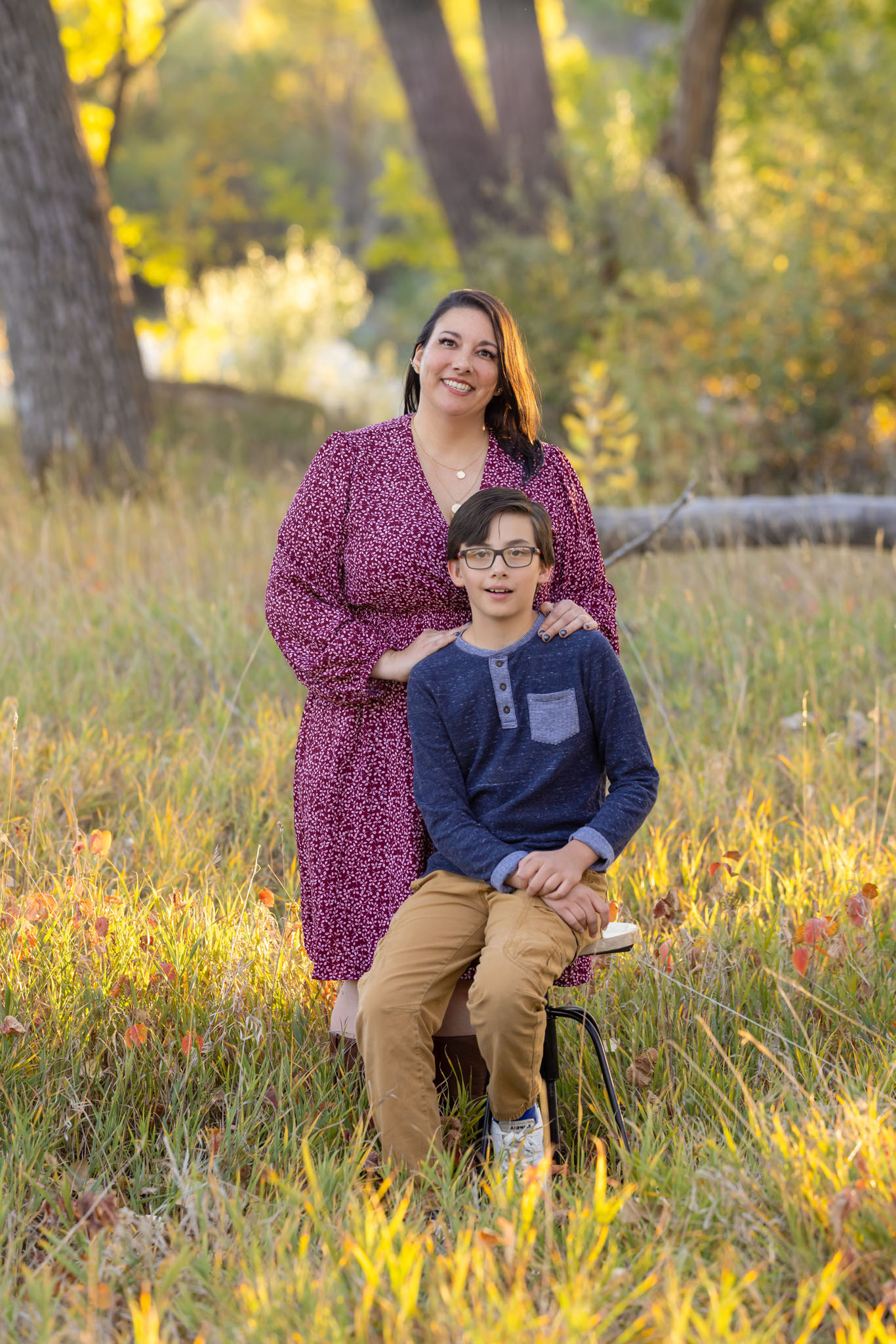 a mom standing behind her middle school aged son who is sitting on a stool with her hands on his shoulders in a clearing in City Park in Pueblo Colorado