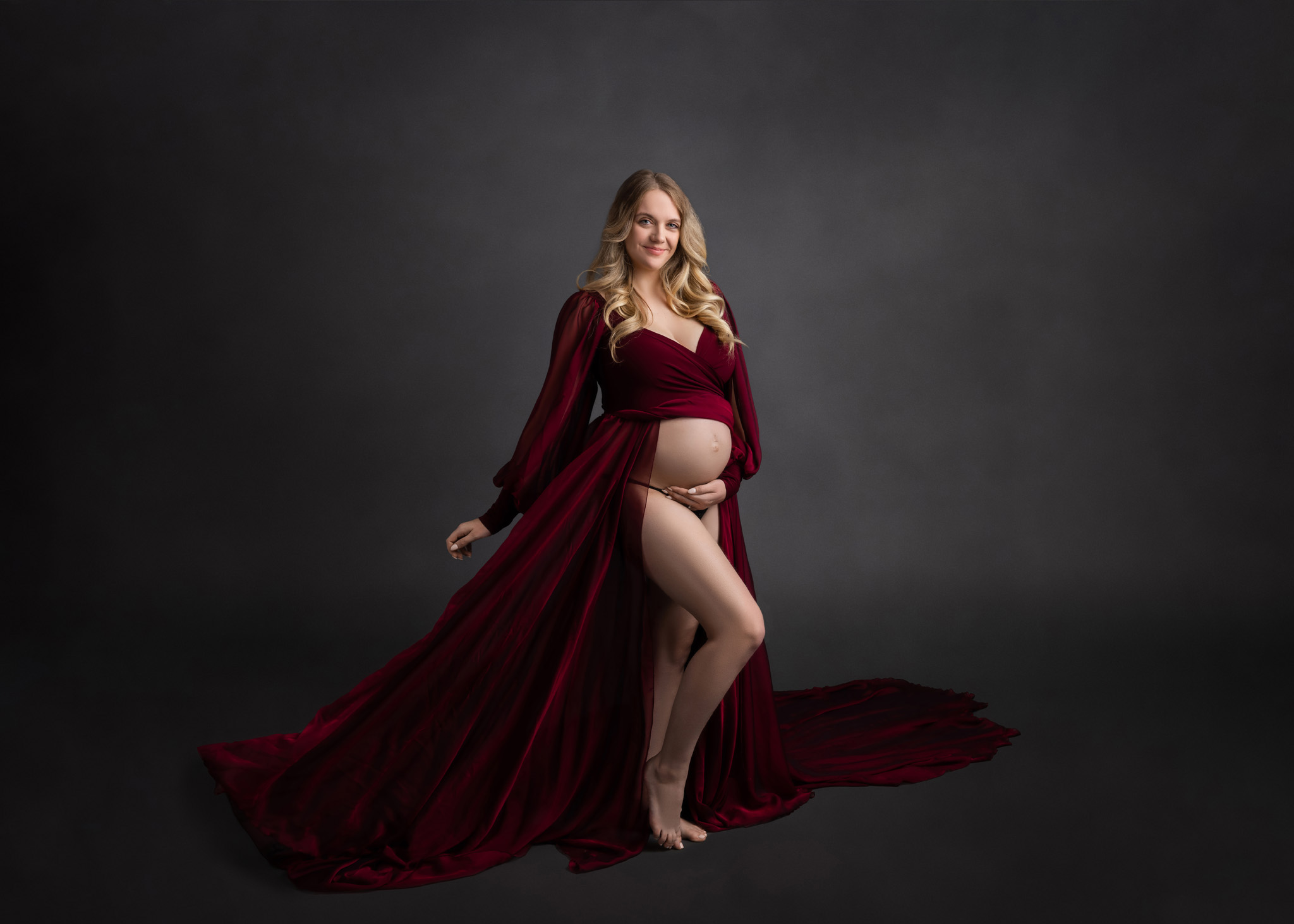 pregnant mom in a flowy maroon dress holding her belly standing in front of a grey backdrop