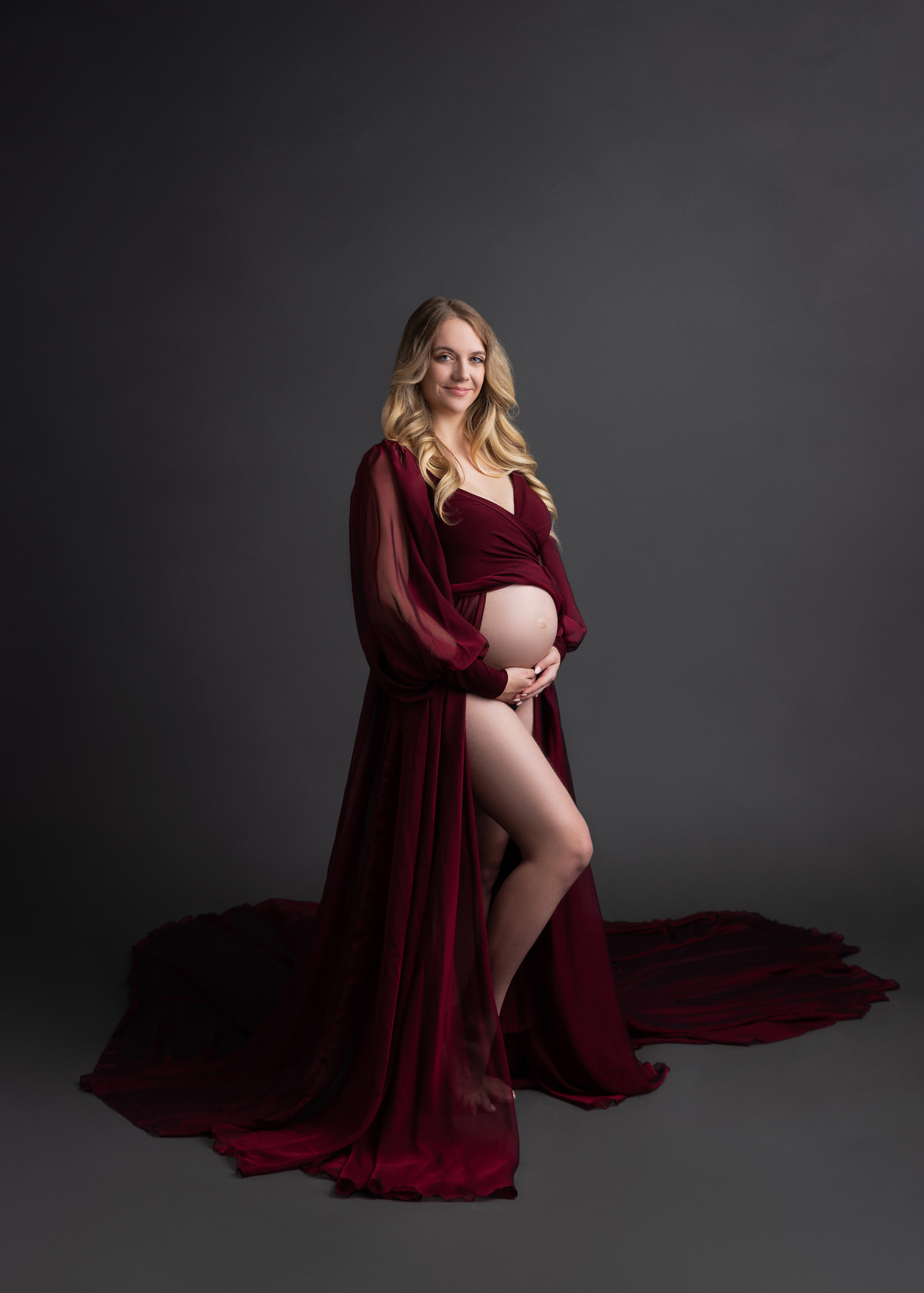 pregnant mom in flowy maroon dress cradling her exposed belly standing in front of a grey backdrop