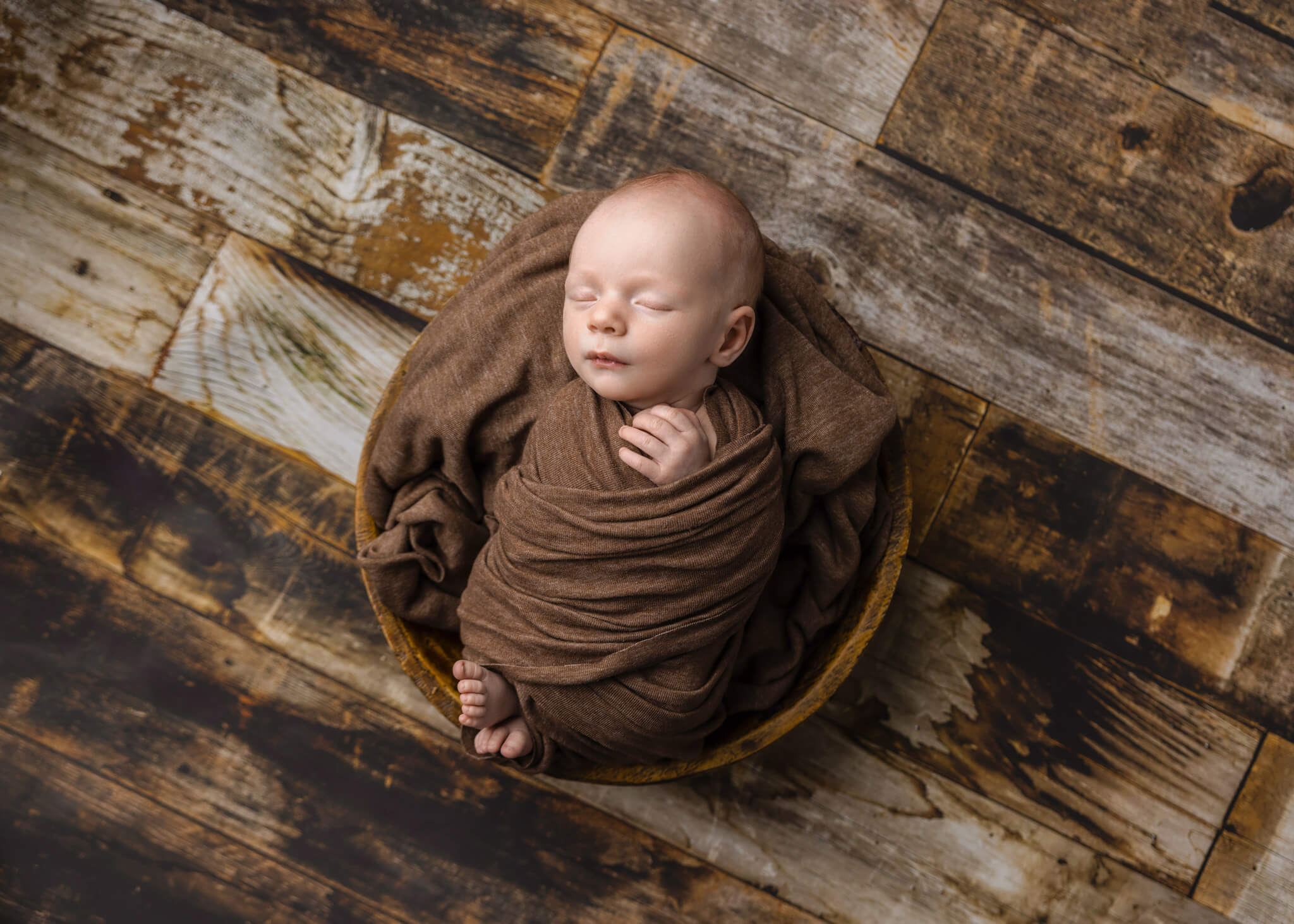 newborn baby in a brown wrap asleep in a small wooden bowl on a wooden floor backdrop