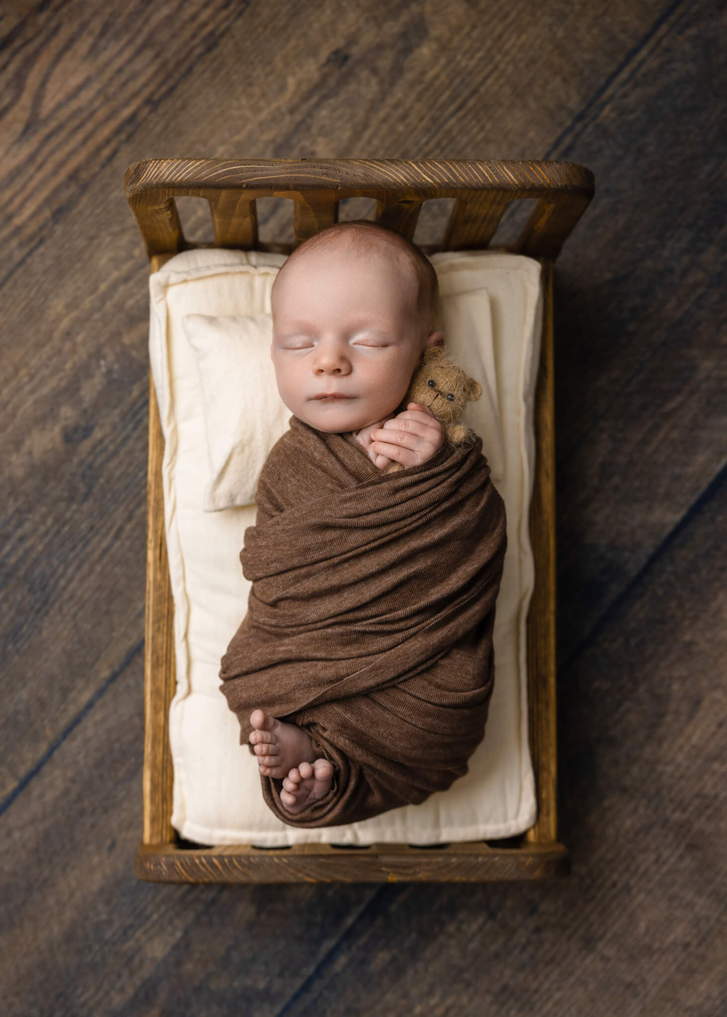 newborn baby asleep in a tiny wooden bed wrapped in a brown blanket holding a tiny teddy bear