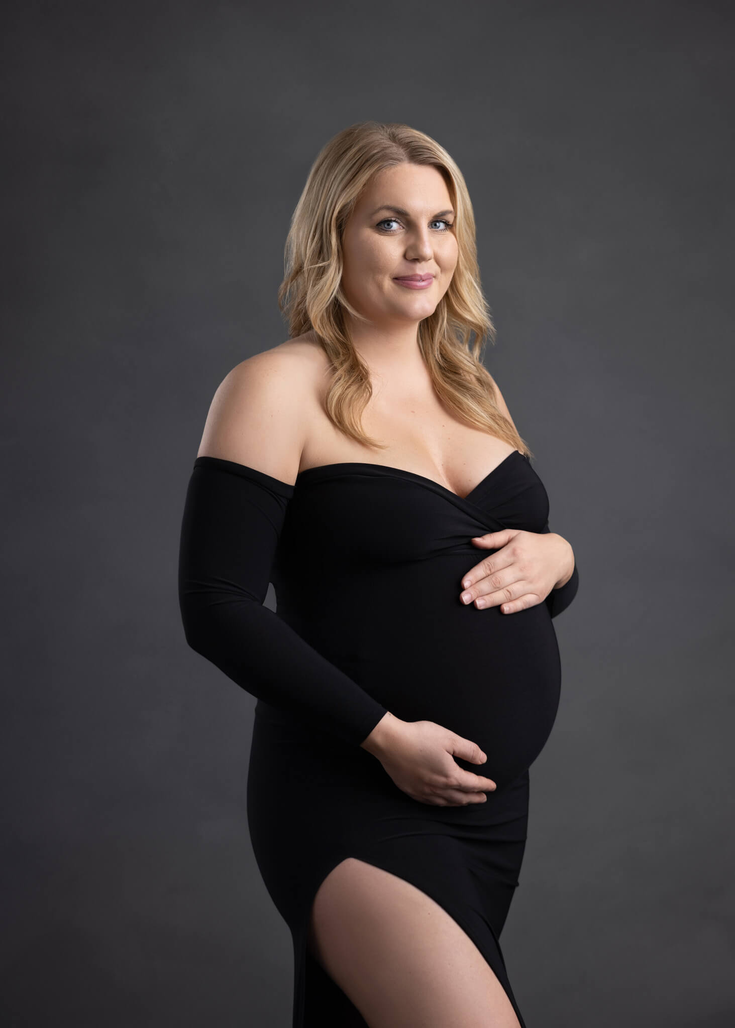 pregnant mom in an off the shoulder black fitted dress cradling her belly standing in front of a grey backdrop