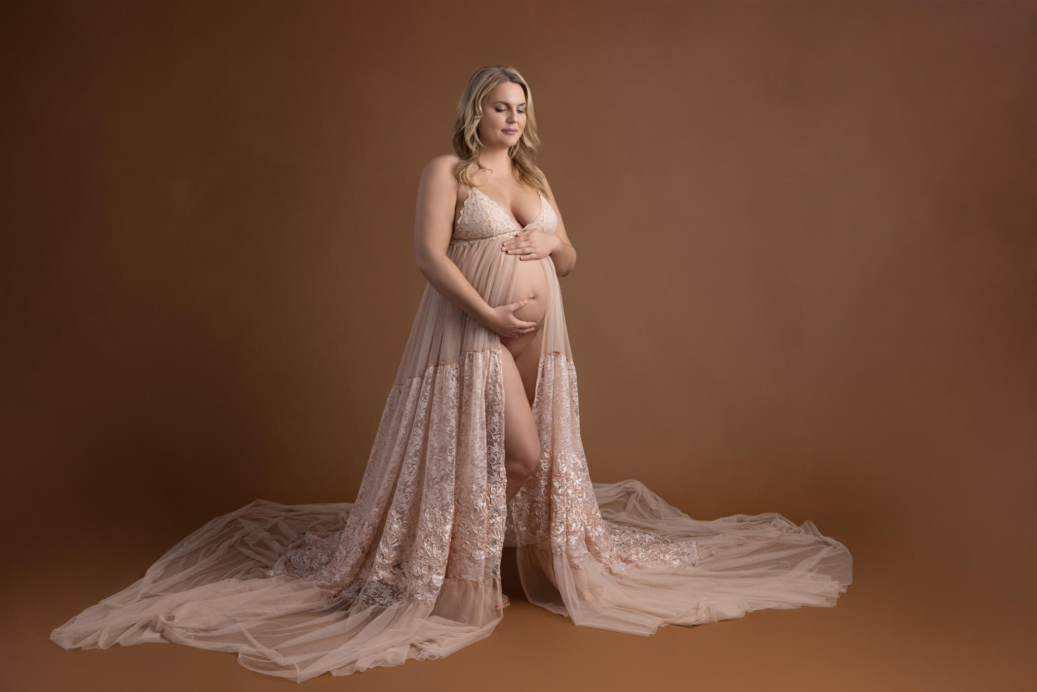 pregnant mom in a flowy cream colored lace dress cradling her stomach standing on a deep cream backdrop