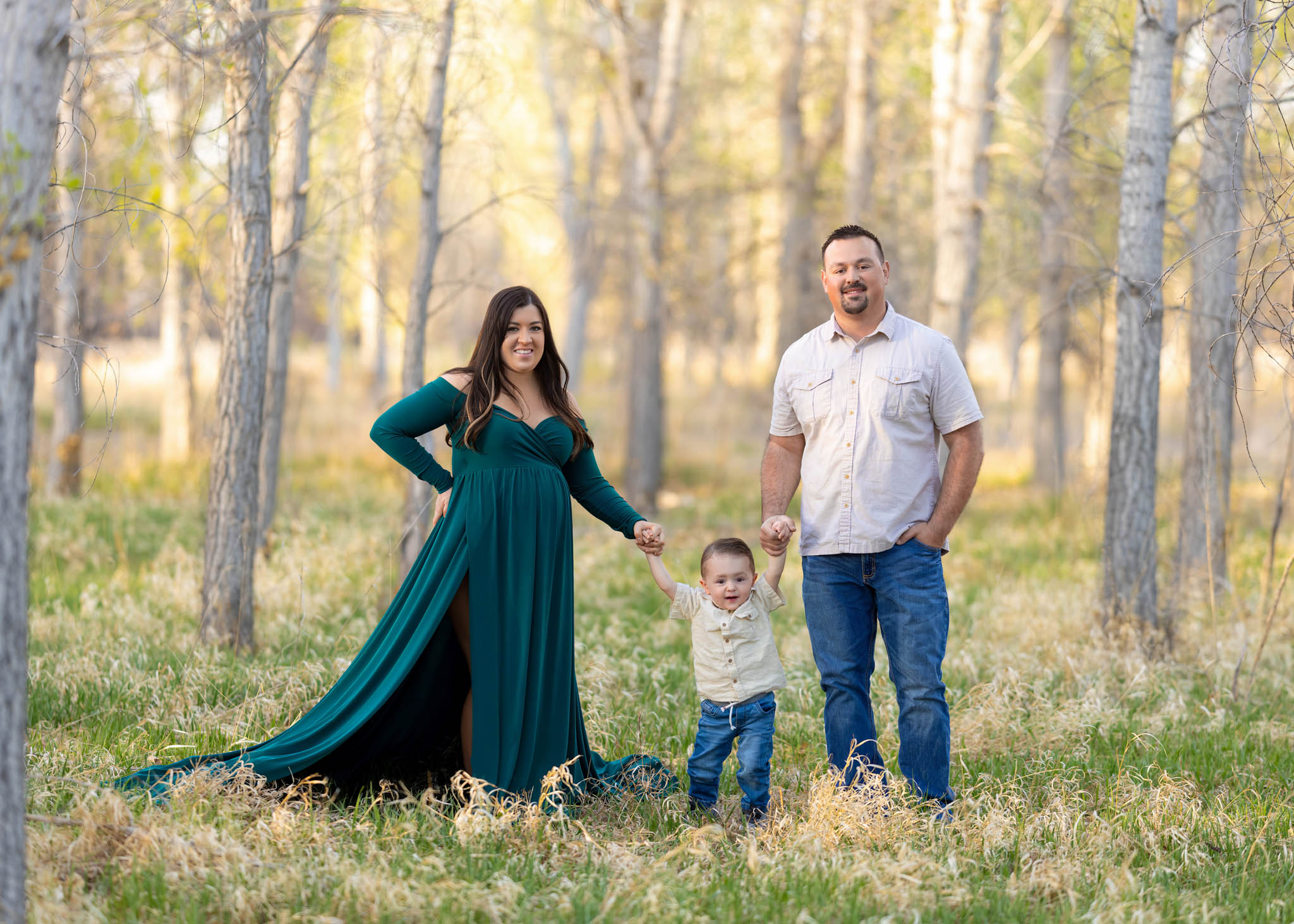 pregnant mom in a flowy green dress holding hands with her toddler son and husband among trees in City Park in Pueblo CO
