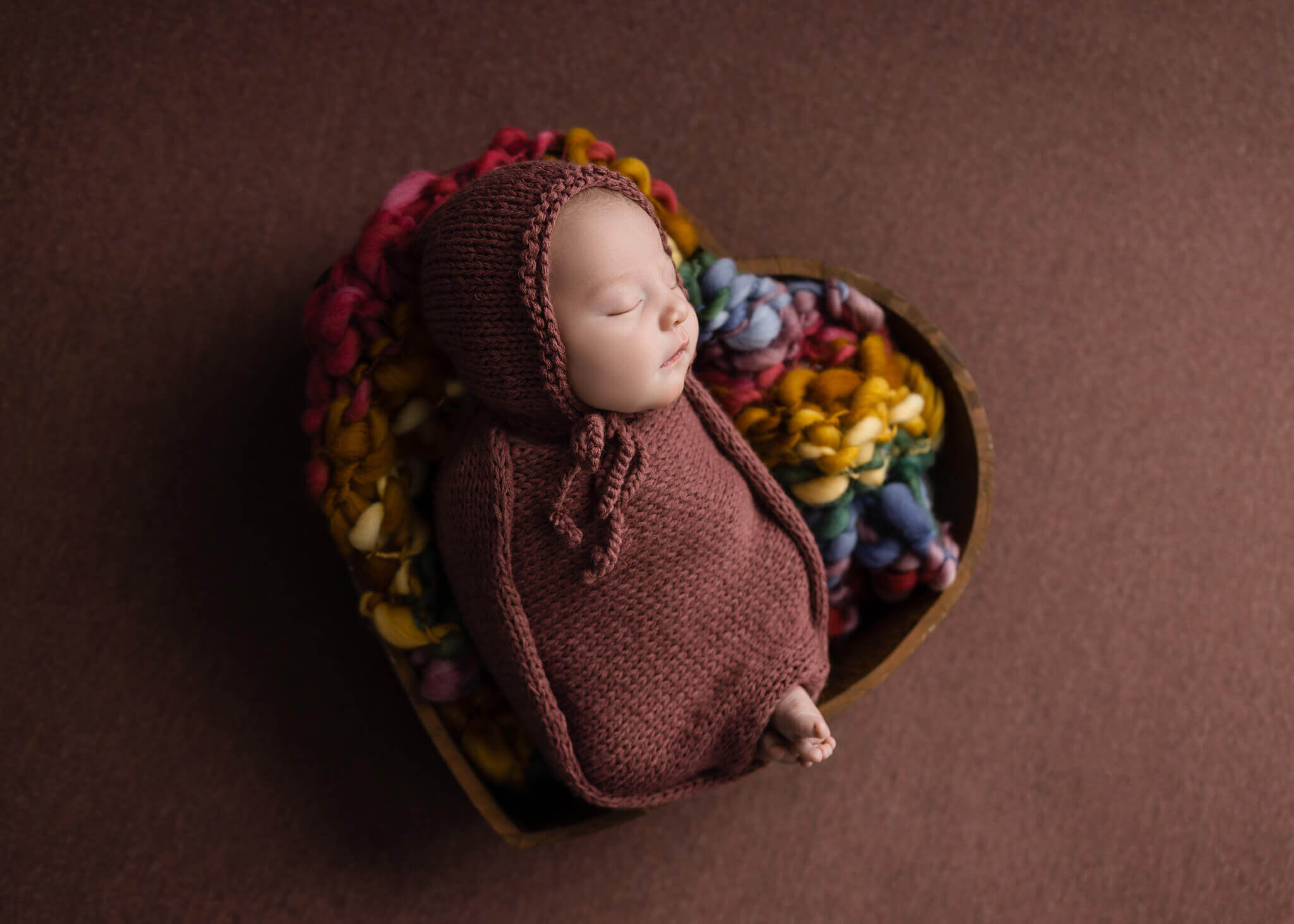 newborn baby asleep on a rainbow blanket in a heart shaped bowl set wrapped in a pink blanket and wearing a pink bonnet
