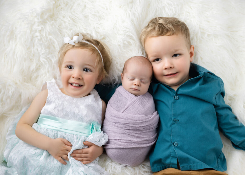 a young brother and sister laying with their newborn sister between them on a white furry rug