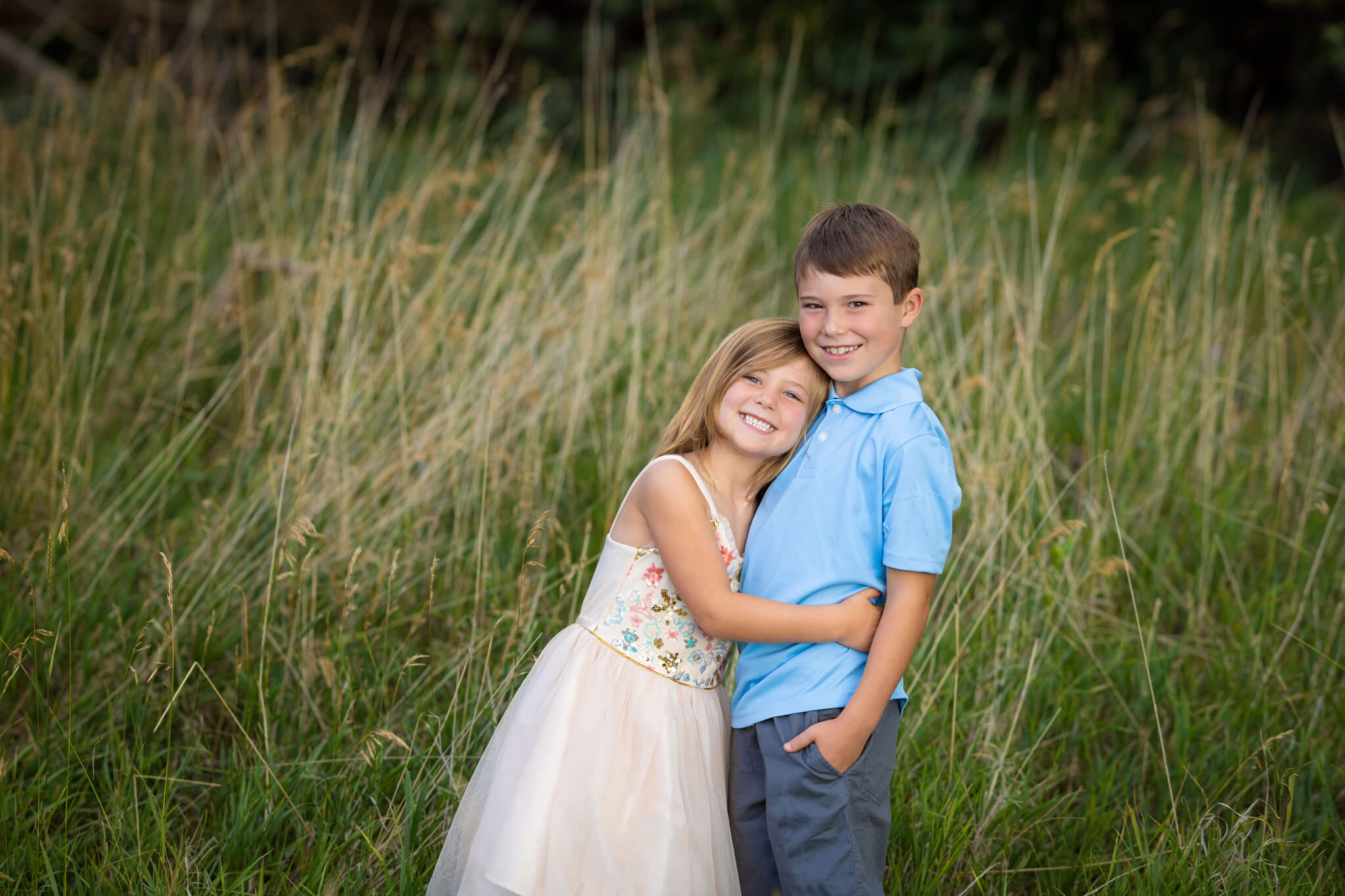 young brother and sister sitting hugging standing in a field of tall green grasses