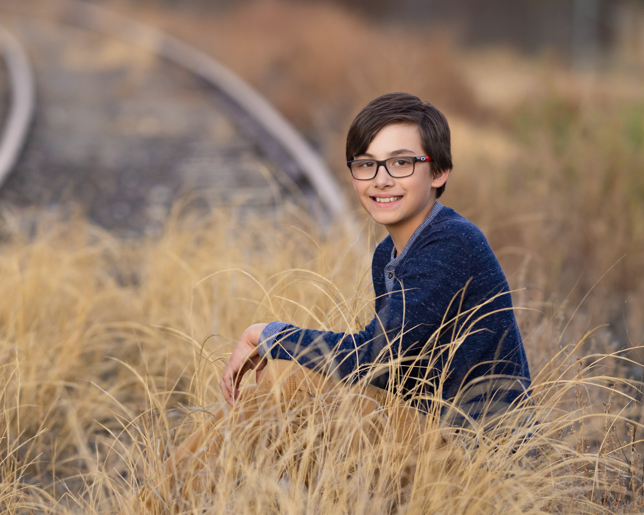 young brown haired boy with glasses sitting in tall brown grass at City Park in Pueblo CO