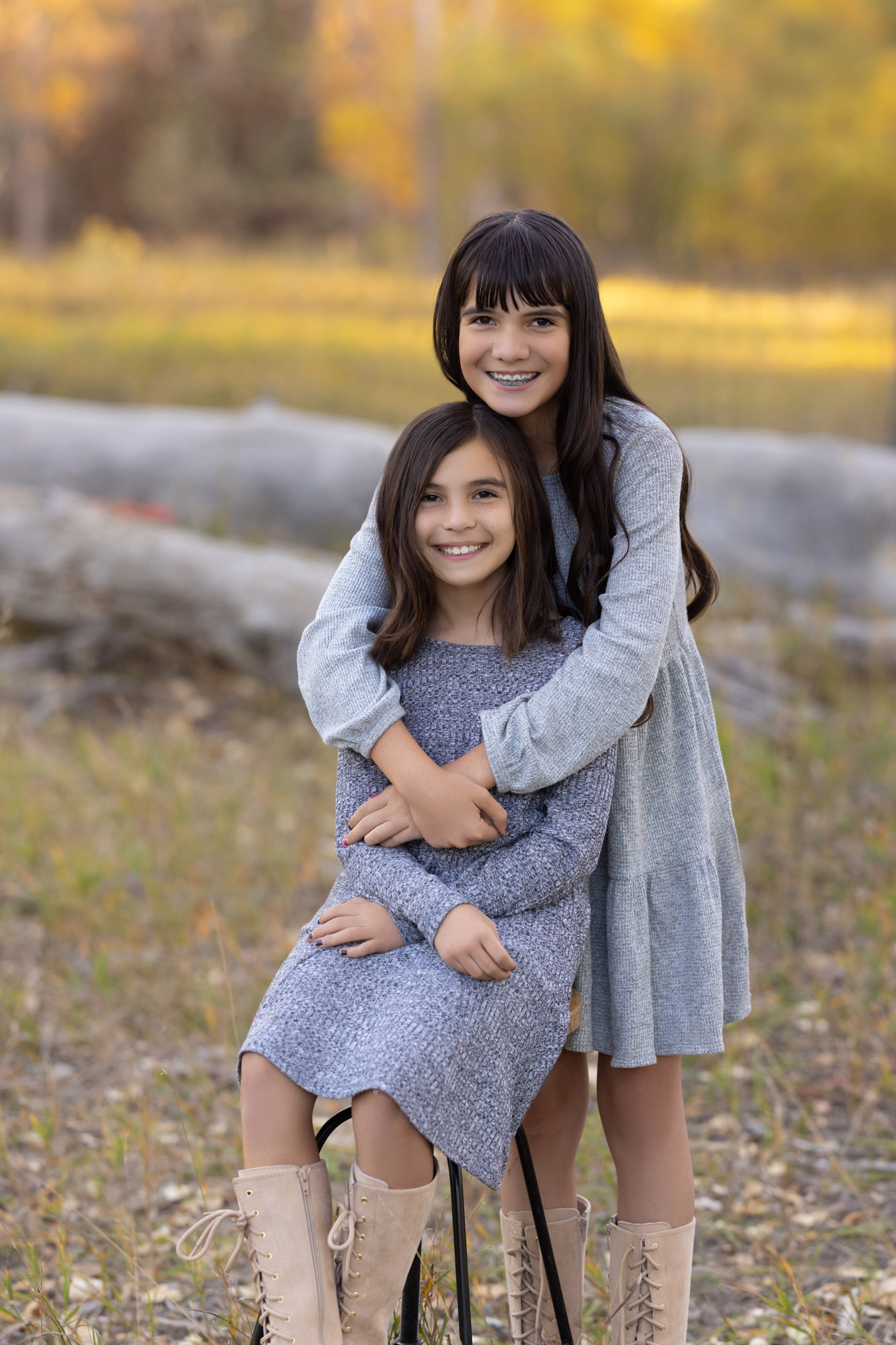 two young sisters, one sitting on a stool and the other standing behind her hugging her, in a field at City Park in Pueblo